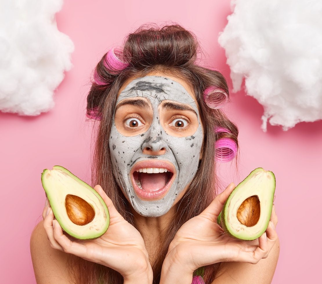 Emotional brunette woman excalims loudly with wide opened mouth applies facial clay mask for skin ca